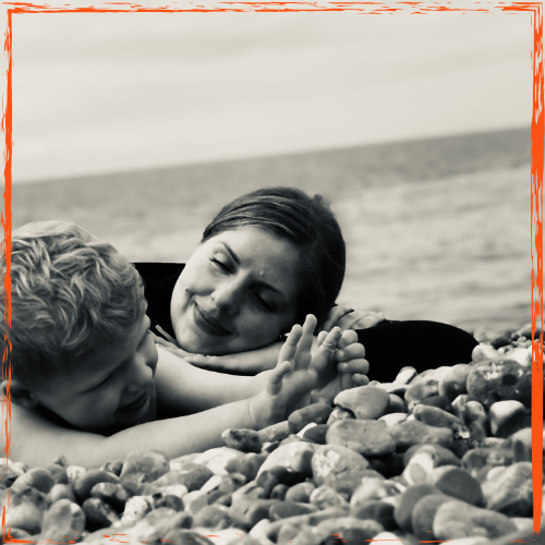 Black and white image of a mother smiling and looking at her small child, both lying down on a beach and playing.